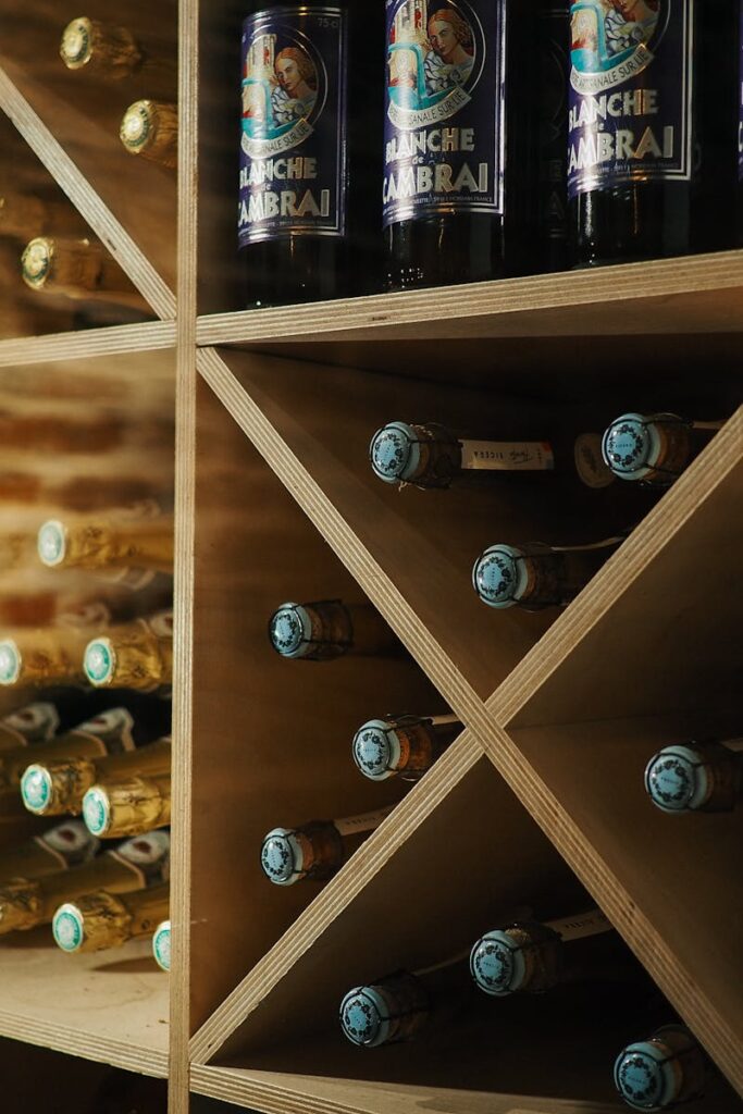 Wooden shelves in a wine cellar showcasing beer and wine bottles with a brick wall backdrop.